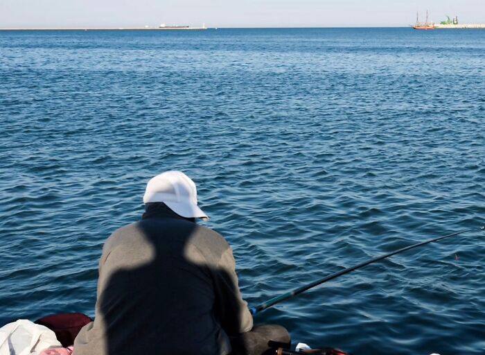 Man in white cap fishing by the sea, captured in a perfectly timed street photo by photographer Melahat Ünel.