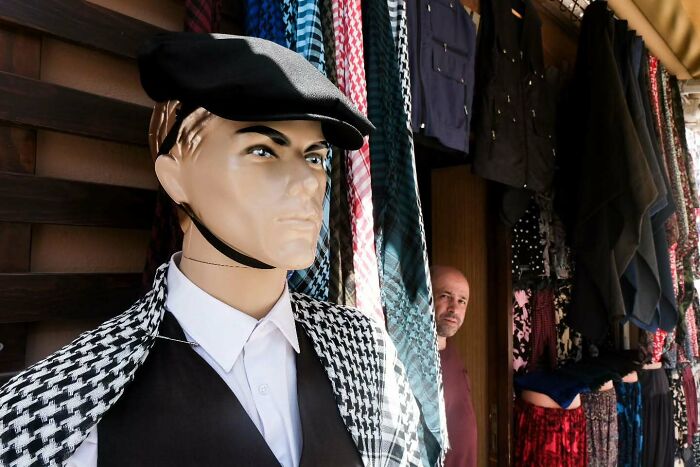 Man peeking from clothing store beside mannequin wearing a hat and checkered shawl in a perfectly timed street photo.