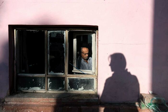 Man sitting by a window with sunlight casting a sharp shadow on a pink wall in perfectly timed street photos.