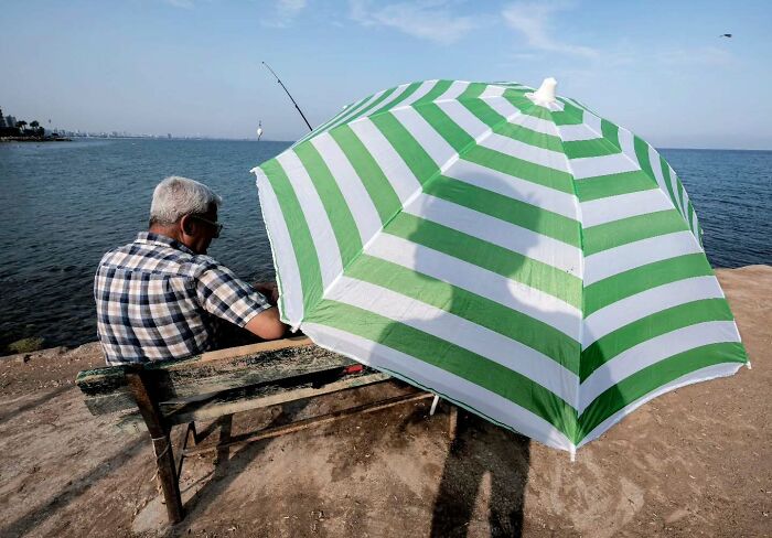 Man fishing by the sea sitting on a bench under a large striped umbrella in a perfectly timed street photo.