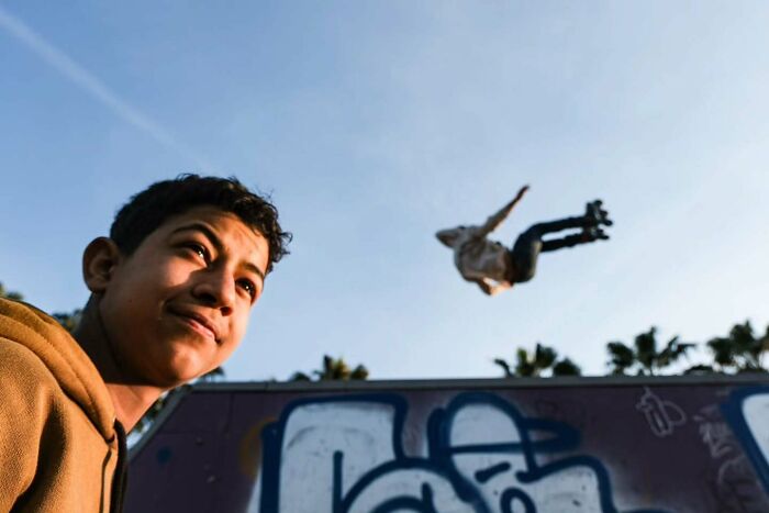Young man watching a skateboarder mid-air at a graffiti-covered skatepark in a perfectly timed street photo.