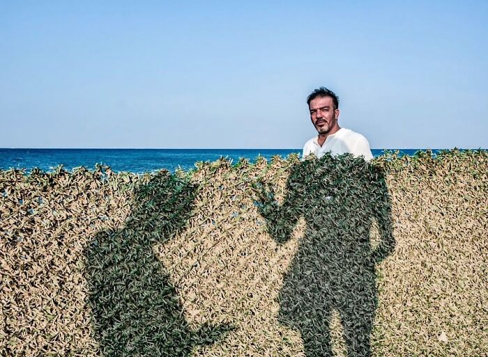 Man standing by the sea with two large shadows on a hedge, captured in a perfectly timed street photo by Melahat Ünel.