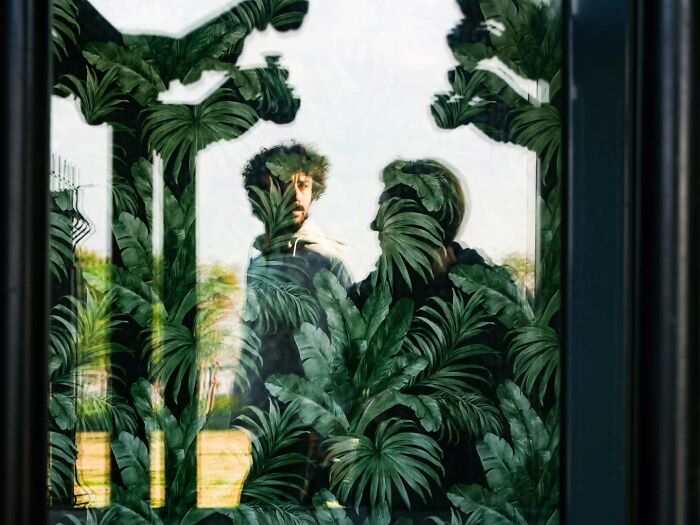 Two men reflected in glass window with tropical leaf patterns, showcasing a perfectly timed street photo moment.