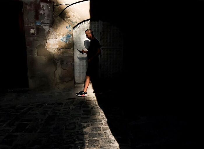 Man walking in a sunlit urban alley, engrossed in his phone, captured in a perfectly timed street photo.