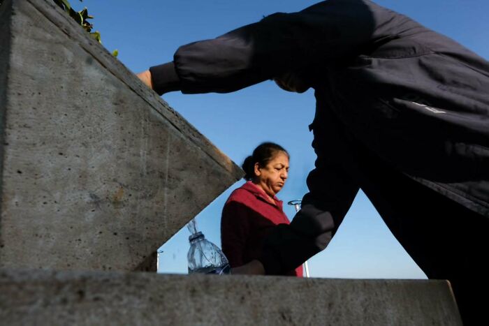Two people interacting near a concrete structure under clear blue sky in a perfectly timed street photo by Melahat Ünel.