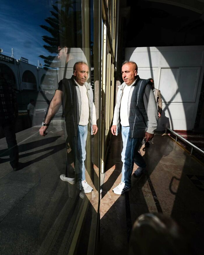 Man walking next to a reflective glass wall creating a mirror effect in a perfectly timed street photo by Melahat Ünel.