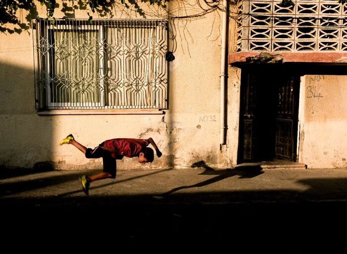 Child captured mid-air jump on street with shadows and textured wall in a perfectly timed street photo by photographer Melahat Ünel.