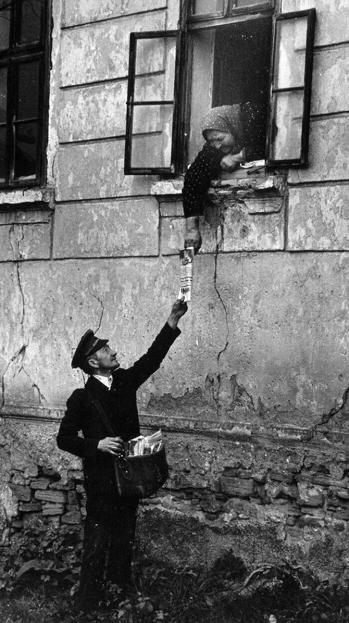 Mail carrier handing newspaper to woman leaning out of window in an old photo showing people’s lives from a different time