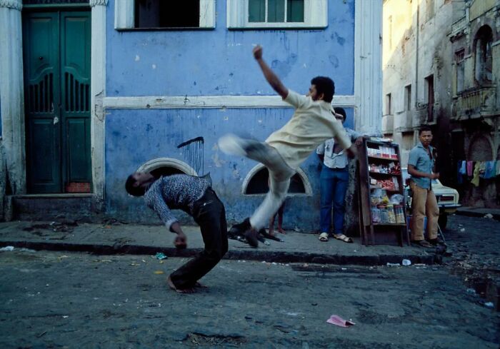 Two men engaged in a street fight in front of a blue building, capturing old photos of people’s lives from a different time.