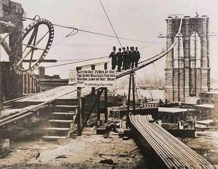Group of men standing on a narrow beam during Brooklyn Bridge construction in an old photo showing people’s lives from a different time.