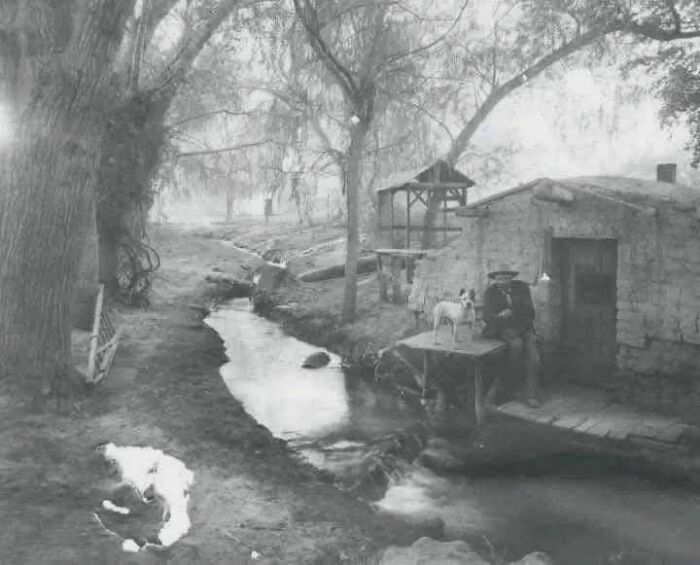 Old photo showing a rural scene with a man, a dog, a small creek, and a rustic house from a different time.