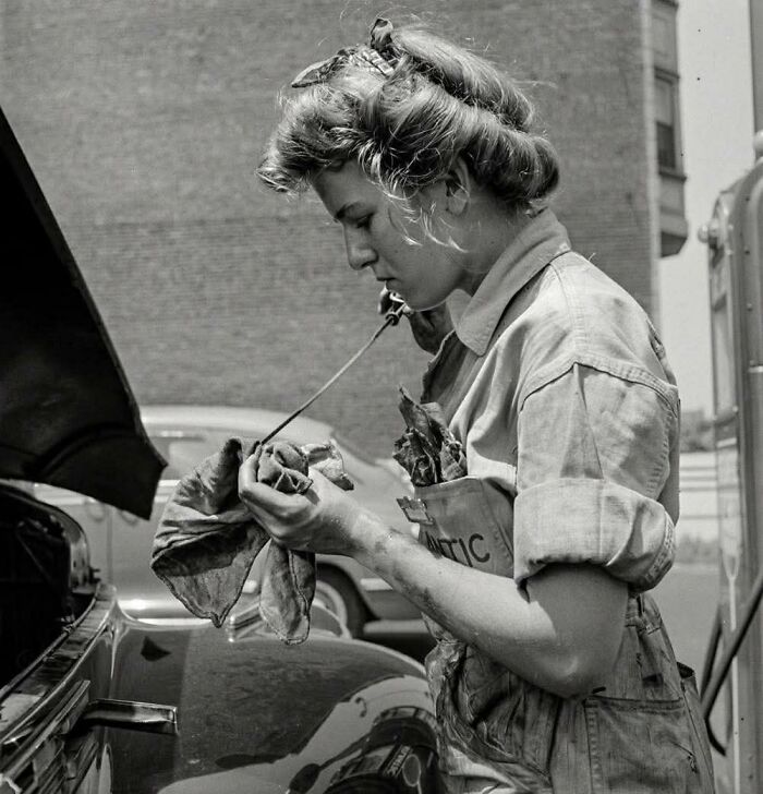 Young woman working on a car engine, wearing a mechanic uniform, capturing old photos of people’s lives from a different time.