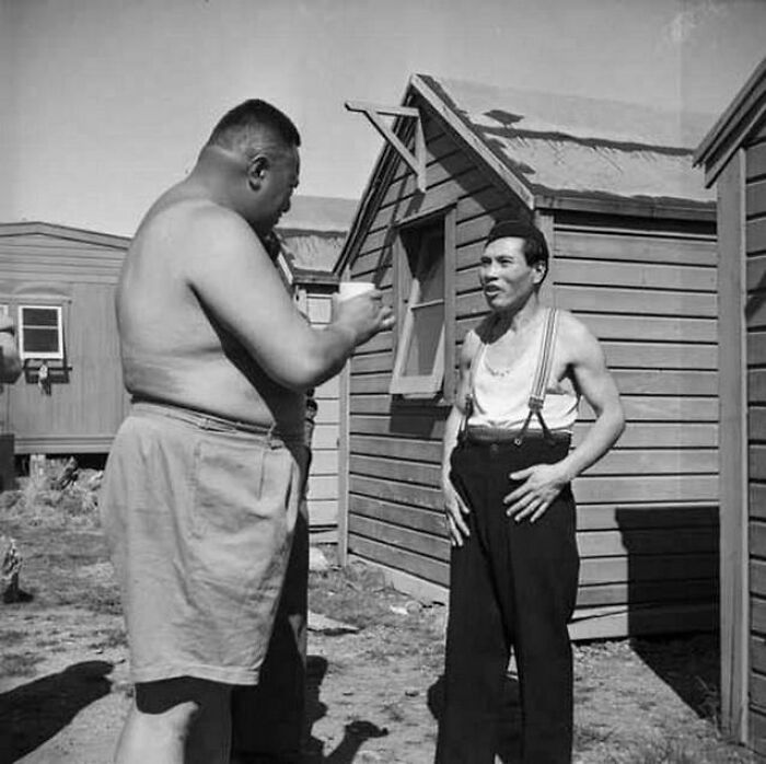 Two men in casual clothes talking outside wooden cabins in an old photo showing people’s lives from a different time.