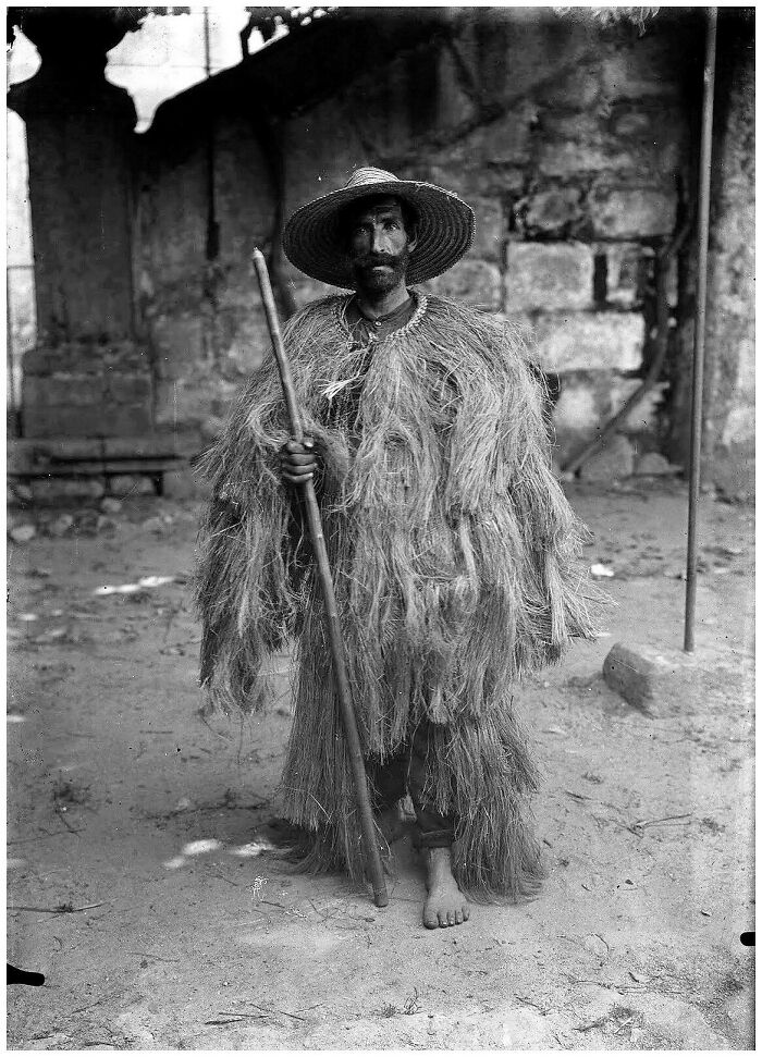 Man in traditional straw outfit and hat holding a staff, old photo showing people’s lives from a different time.