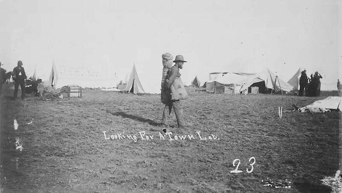 Old photo showing people’s lives from a different time with tents and individuals walking on a grassy field.