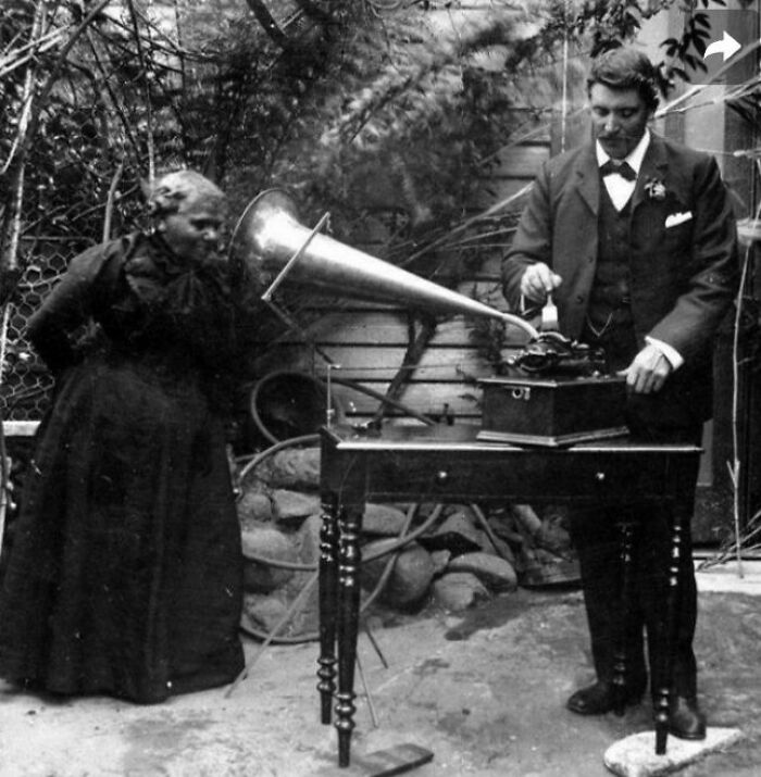 Black and white photo showing people from a different time using an early phonograph with a large horn speaker outdoors.