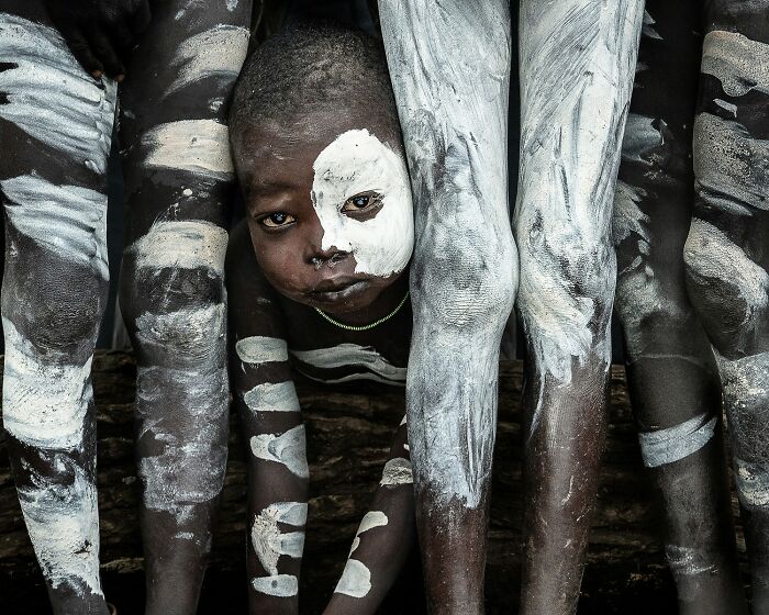 Child with white face paint peeking through legs painted in white and black patterns, reflecting Earth's hidden patterns in photography.