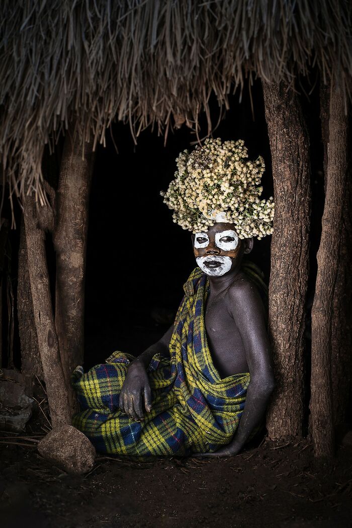 Person with floral headdress and white face paint sitting wrapped in colorful cloth, captured in stunning aerial color photography.