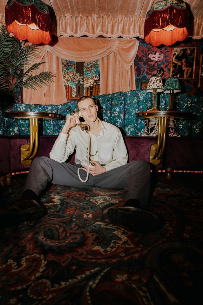 Young man sitting on patterned carpet in vintage room, surrounded by ornate decor and colorful upholstery, holding telephone receiver