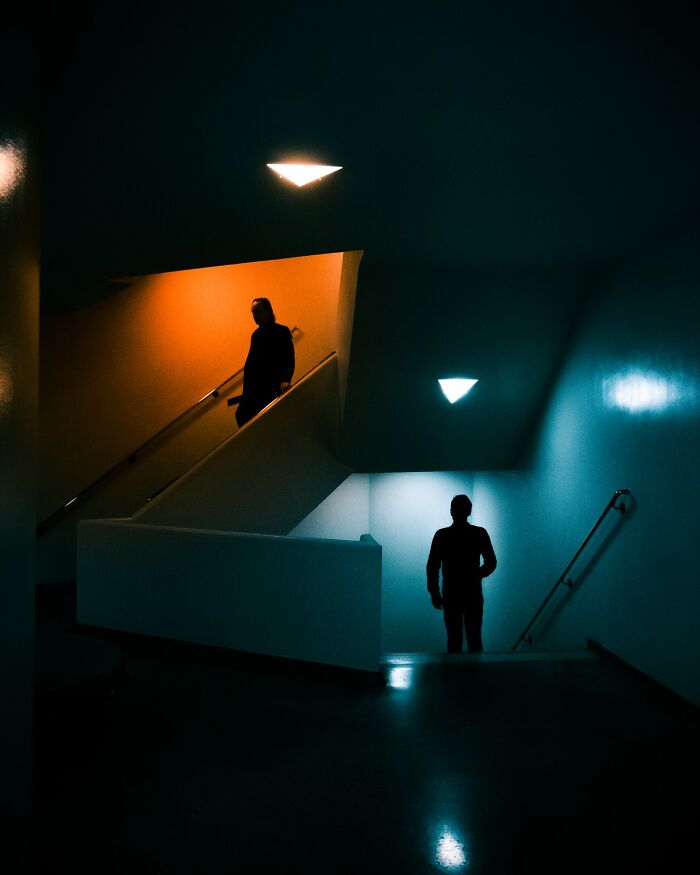 Dark stairwell with two silhouetted figures illuminated by contrasting warm and cool lights, showcasing hidden patterns and color photography.