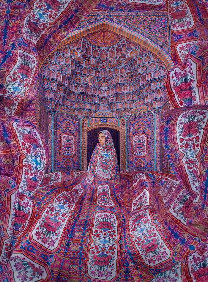Woman wrapped in vibrant, patterned fabric standing in a room filled with intricate aerial color photography patterns.