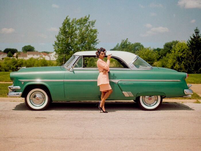Woman in pink outfit with hair rollers standing beside a vintage green car on a sunny day outdoors.