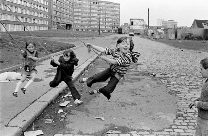 Children playing on swings in an urban setting, captured in an old photo showing people's lives from a different time.