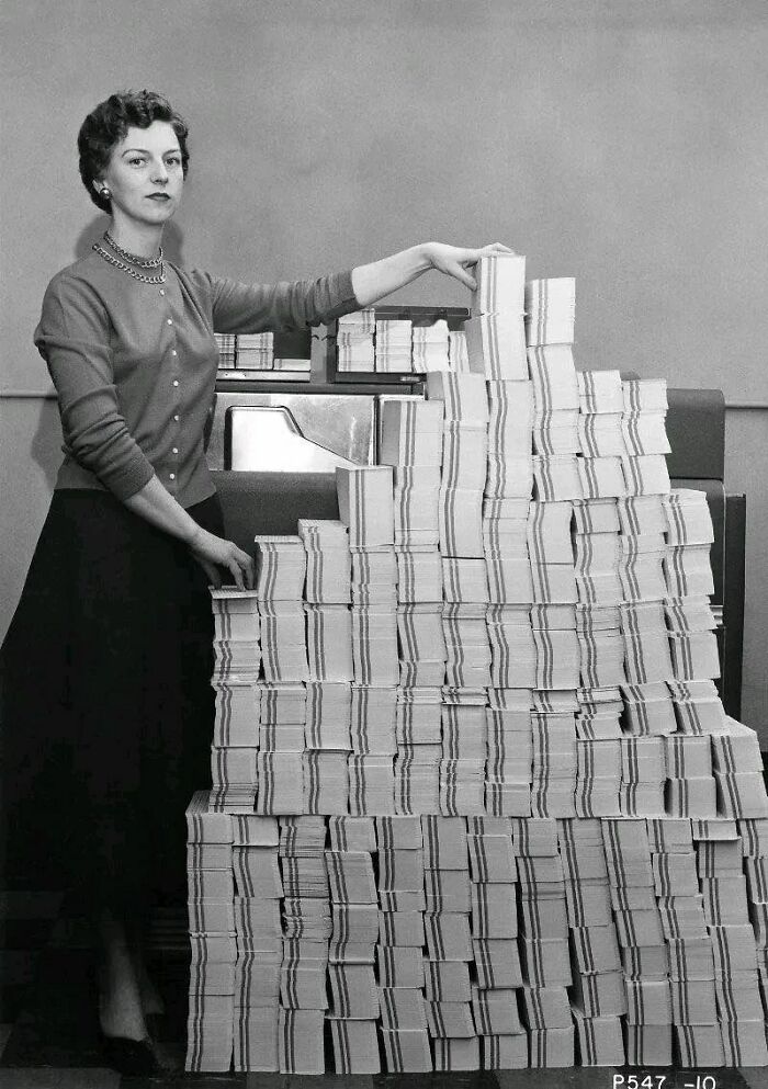 Woman standing beside large stacks of paper, illustrating old photos showing people’s lives from a different time.