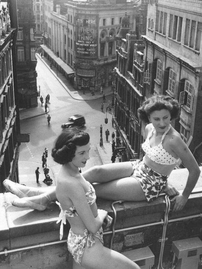Two women in vintage swimsuits sitting on a rooftop ledge overlooking a busy city street in old photos of people's lives.