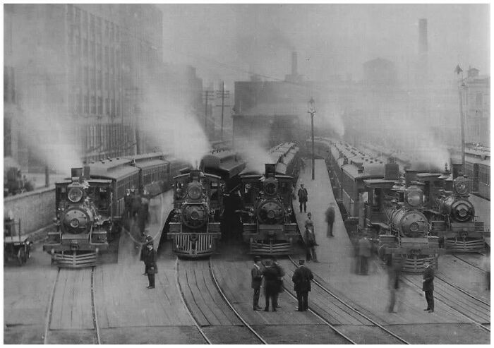 Old black and white photo of steam trains and people at a busy station showing lives from a different time period.