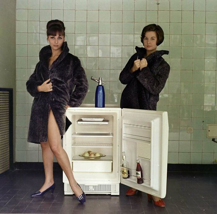 Two women in fur coats posing beside an open refrigerator, illustrating people’s lives from a different time in old photos.