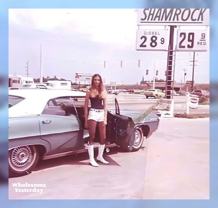 Young woman in 1970s fashion standing beside a vintage car at a gas station, showing people’s lives from a different time.