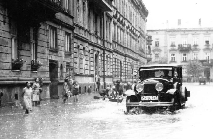 Vintage photo of an old car driving through a flooded street with people walking and watching alongside historic buildings.