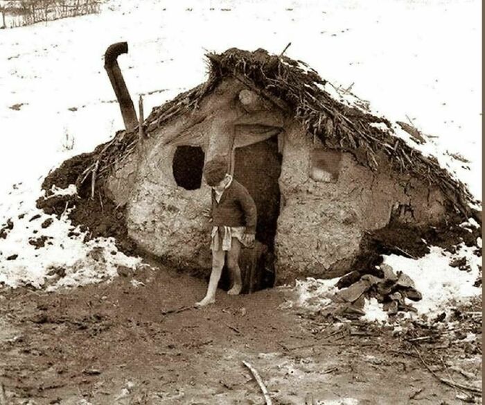 Man walking barefoot outside a small rustic shelter in winter, showcasing old photos of people's lives from a different time.