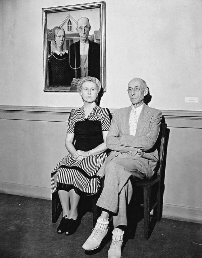 Black and white photo of an elderly couple seated under a famous painting, capturing people’s lives from a different time.