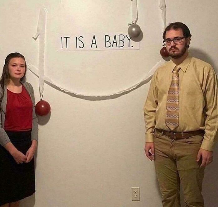 Couple standing by a wall with balloons and a sign reading "It is a baby" in a funny and bizarre image combining the holy and cursed.