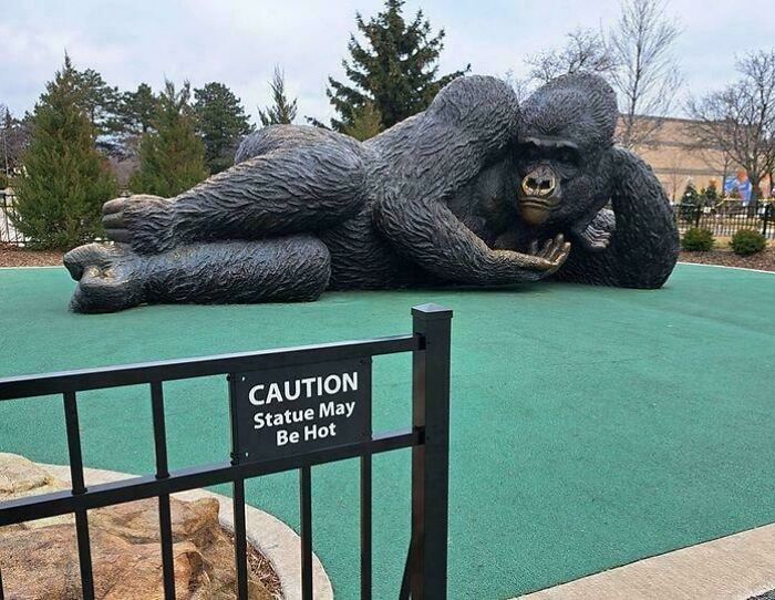 Large reclining gorilla statue in a park with a caution sign, a funny and bizarre image combining the holy and cursed concept.