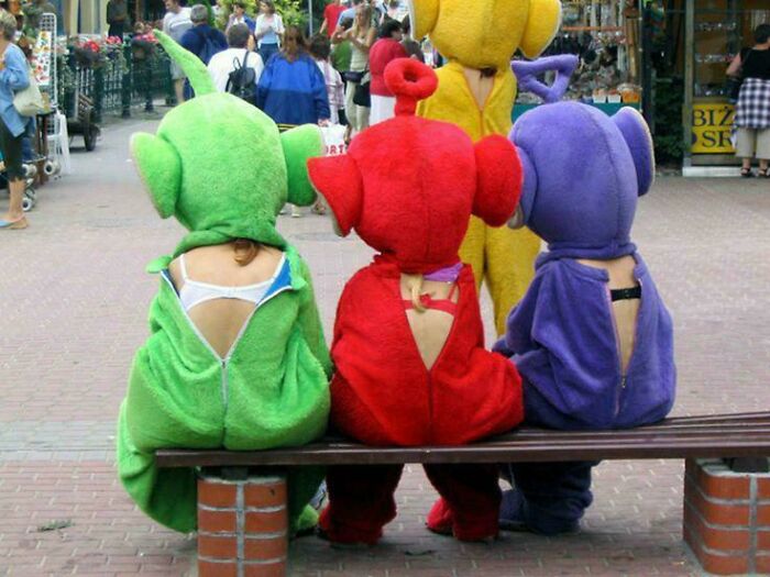 Three people wearing colorful Teletubby costumes with broken back zippers sitting on a bench in a busy street.