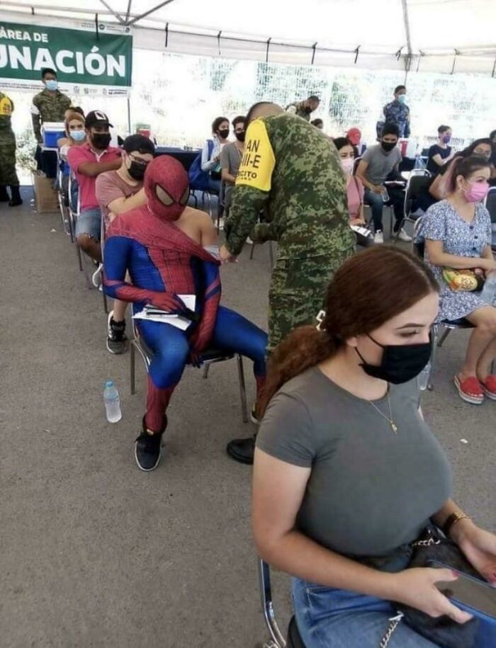 Person dressed as Spider-Man sitting and receiving a vaccine shot from a soldier in a crowded vaccination area.