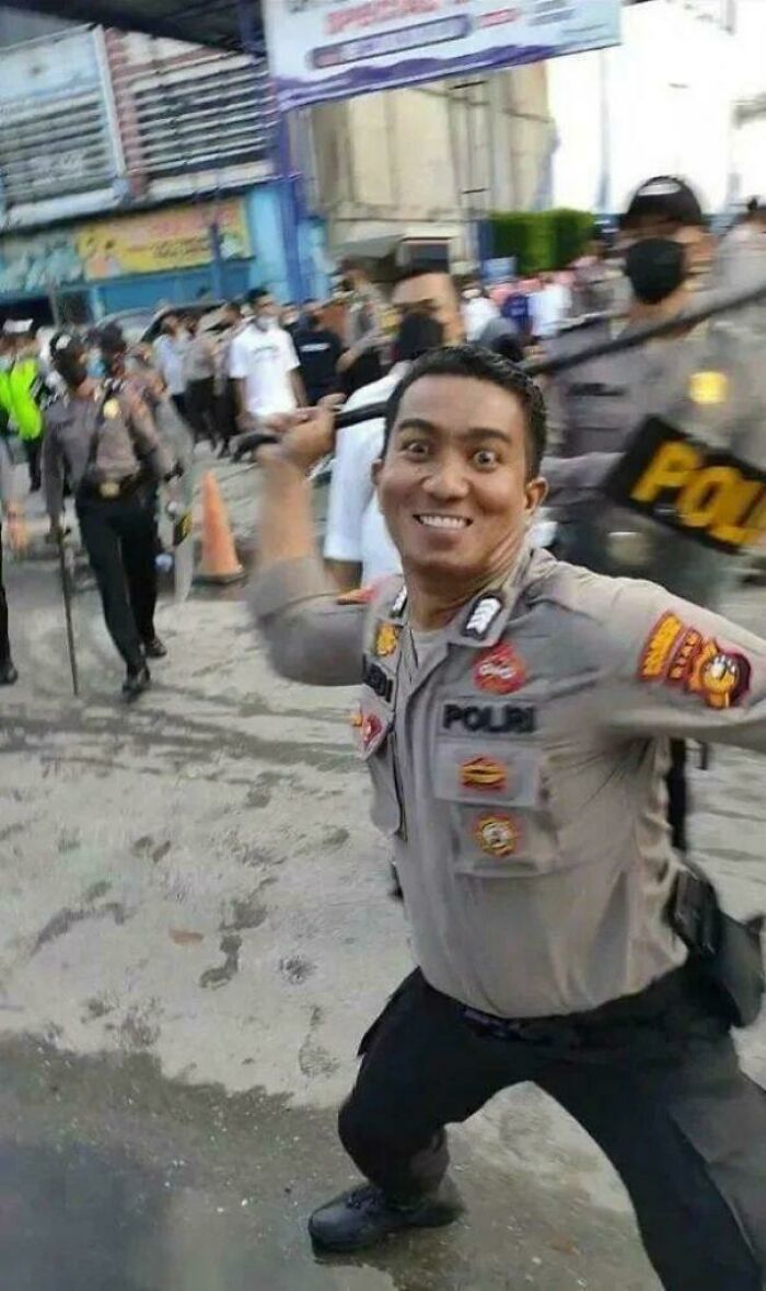 Smiling police officer in uniform holding baton in a crowded street, capturing a funny and bizarre moment.