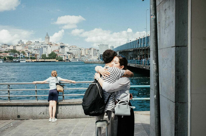 Couple embracing near waterfront with city and bridge in background, capturing candid life moments in Turkey by Yusuf Aksoy.