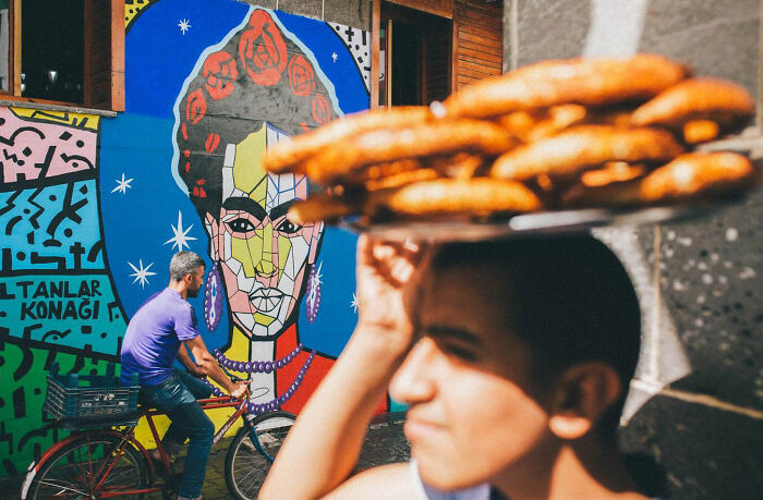 Man carrying simit bread on head with colorful street art and cyclist in the background, capturing candid life in Turkey.