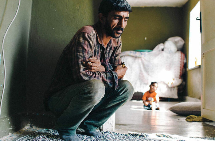 Man crouching in a modest room with a child in the background, capturing candid life in Turkey by Yusuf Aksoy.