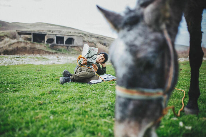 Child reading a book while lying on grass near a donkey, showcasing candid life moments in Turkey by Yusuf Aksoy.