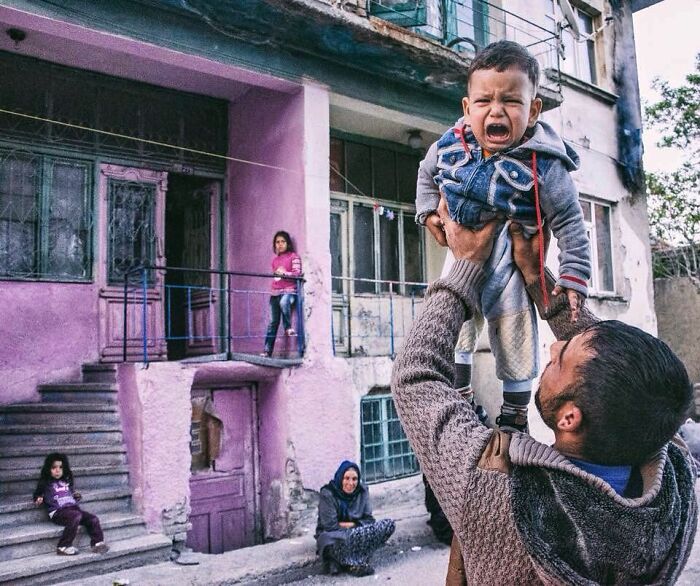Candid photo of a man lifting a crying child outside a weathered building capturing real life in Turkey by Yusuf Aksoy