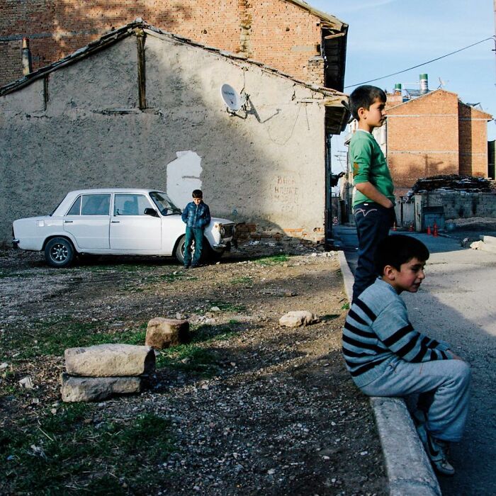 Three boys in a rural Turkish neighborhood with an old car and weathered brick buildings, showcasing candid life in Turkey.