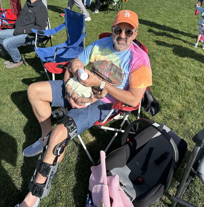 Man with leg brace wearing a tie-dye shirt and orange cap feeding a baby doll, showcasing one of the funniest parents ever.