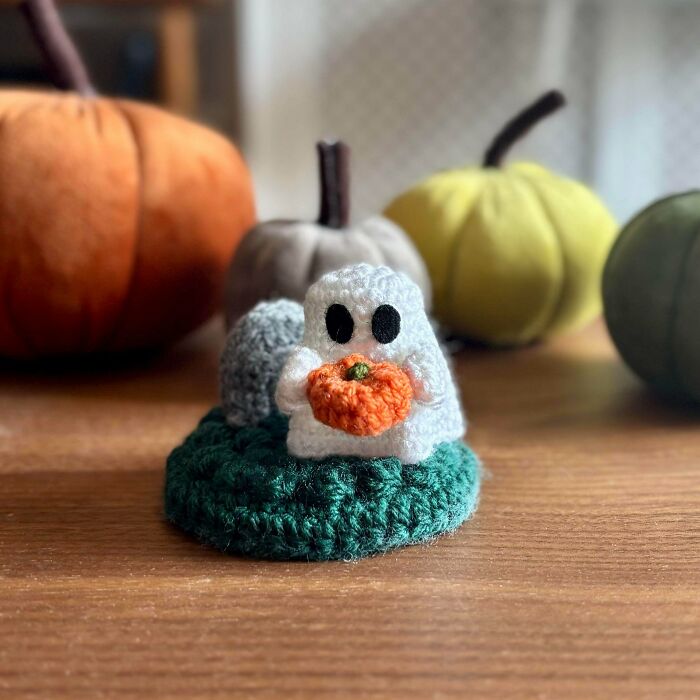 Crocheted ghost holding a small pumpkin on a green base with fabric pumpkins blurred in the background.