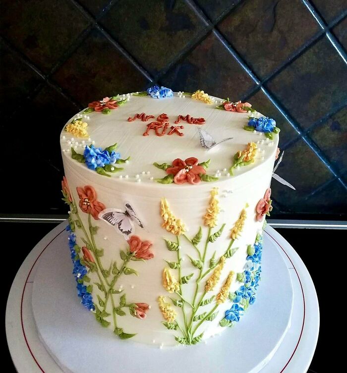Tall white cake decorated with colorful floral piping and butterfly designs, showcasing gorgeous baked goods artistry.