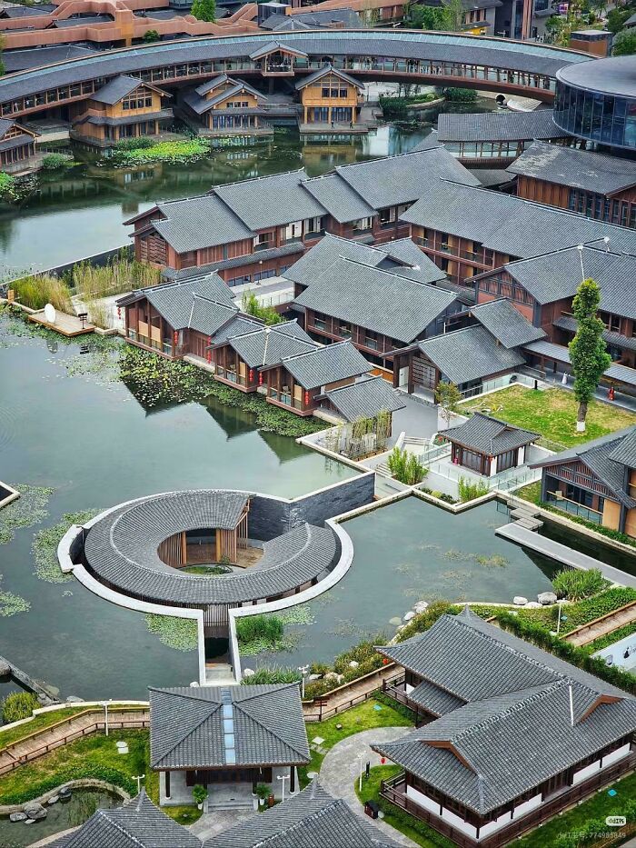 An aerial view of beautiful architecture of a village built on and around water, with traditional roofs and a circular structure in the pond.